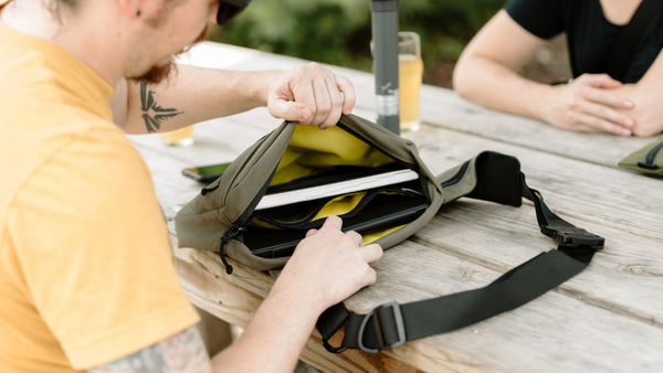 The city sling bag is an everyday carry bag great for notebooks, ipad and accessories.  Man sitting outside at a picnic table showing the inside of the City Sling Bag.