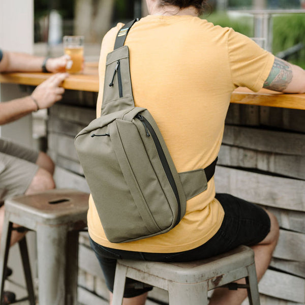 Man wearing ranger green city sling bag sitting at an outside bar talking with a friend